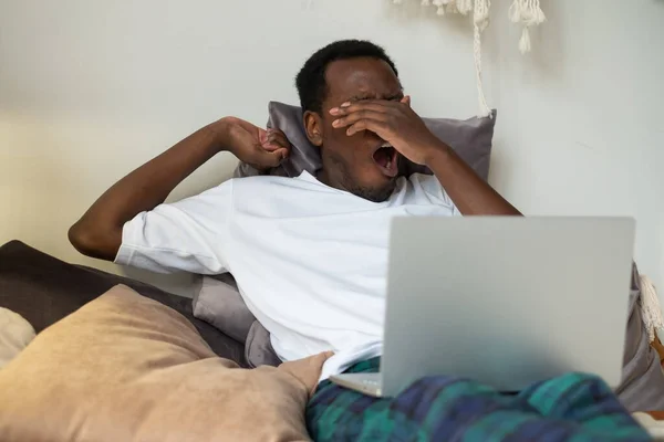 Africano homem trabalhando deitado na cama em casa estar cansado bocejo. — Fotografia de Stock