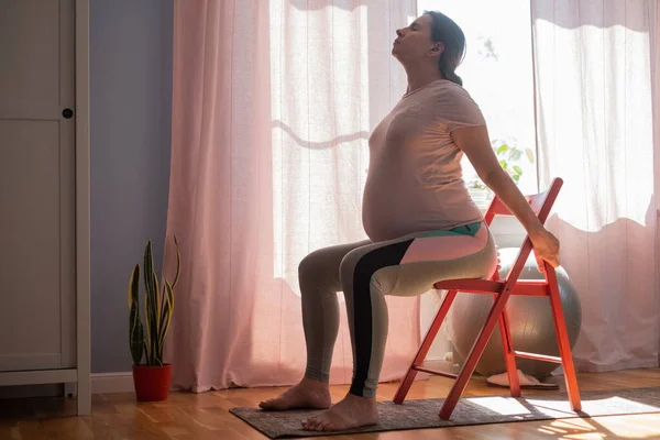 Mulher grávida trabalhando fora dentro de casa praticando ioga em casa usando cadeira — Fotografia de Stock