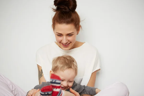 Young mother with her one years old little son dressed in pajamas are relaxing and playing in the bedroom at the weekend together, lazy morning, warm and cozy scene. Pastel colors, selective focus.