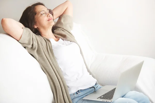 Portrait of attractive middle-aged brunette businesswoman in eyeglasses at home working on laptop computer while work at office work. Content woman sitting on her couch using laptop smiling in room
