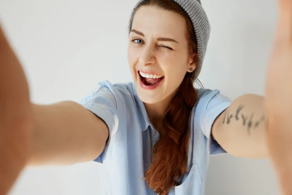 Wide-angle shot of funny young woman having fun with her friends during weekend. Portrait of stylish tattooed girl winking at the camera with mouth wide open. Human face expressions and emotions