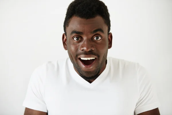 Close up of amazed young African American man in white blank T-shirt, having fun indoor, looking at the camera with excited expression, astonished with sale prices. Human face expressions and emotions