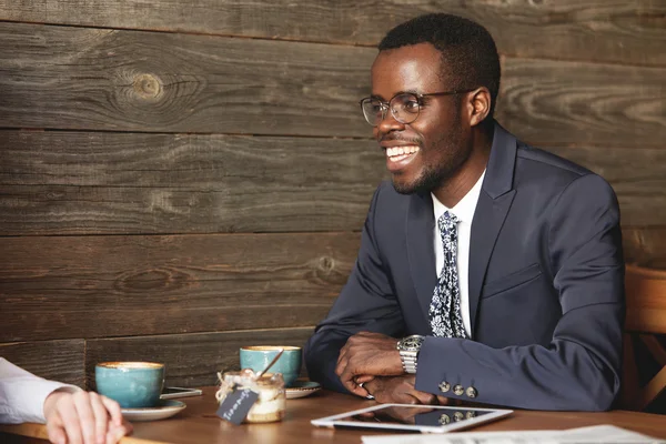 African businessman in formal suit smiling to his job partner discussing collaboration and profit opportunities during coffee meeting at cafe. Entrepreneur has tablet with wireless connection.