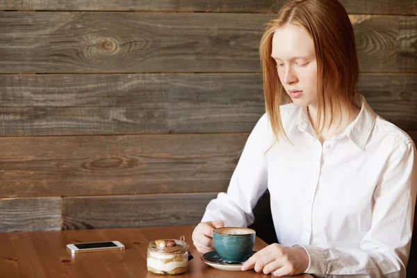 Student girl with ginger hair sitting at the table at a cafe, enjoying coffee and eating dessert after classes at college. Young female office worker relaxing at a coffee shop after hard day at work