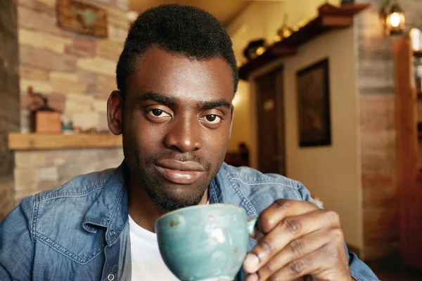 Selective focus. Headshot of young dark-skinned student having a cup of strong espresso after sleepless night he spent, preparing for exams while sitting against the background of a coffee shop