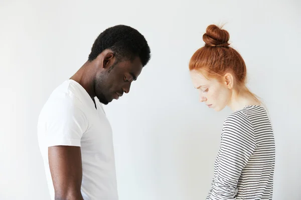 Sad couple looking down with their heads bowed in front of each other. Side view portrait of two sorrowful people: young Caucasian redhead girl and Afro-American melancholic man. Negative emotions.