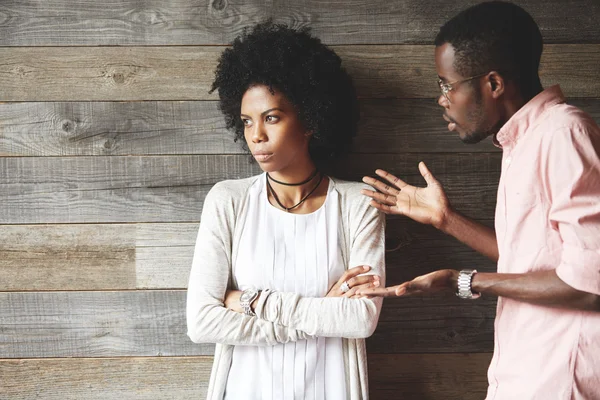 Young black indignant male gesticulating, arguing with his dark skinned girlfriend, who is standing against wooden wall with crossed arms, looking away with deeply offended expression on her face — Stock Photo