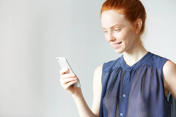 Charming attractive white student girl wearing blue stylish spotted dress skyping with her friend using mobile phone, smiling, looking happy and shy, having healthy freckled skin and tender smile