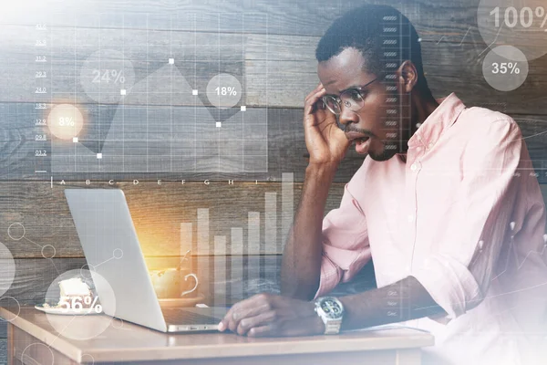 Double exposure of African American entrepreneur looking at laptop screen with surprised expression, shocked with some unexpected news related to his business, with mouth wide open, touching his face