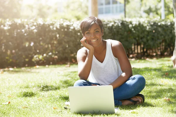 Young successful African American woman blogger sitting in the park on the grass, writing a new post for her popular blog on spcial networks using notebook, looking happy, resting her chin on the hand
