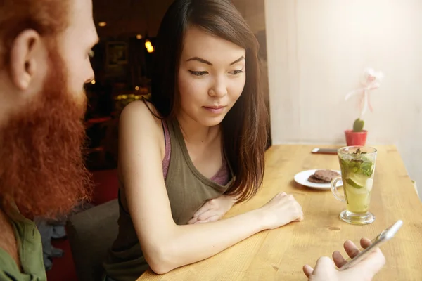 Two young colleagues sitting at cafe during lunch break, drinking flavored tea, having nice conversation. Food blogger holding cell phone, showing his female companion his post in social network
