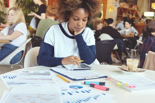 Young African woman entrepreneur with serious concentrated expression sitting at coworking cafe with touch pad pc and papers, analyzing financial information on tablet, resting elbow on table