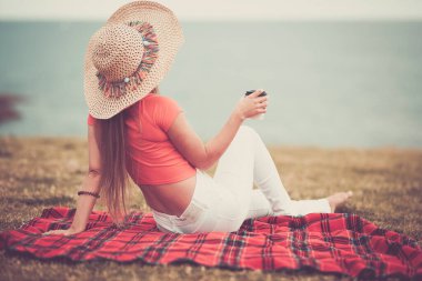 Arkadan seyreden moda genç bayan rahatla ve okyanus manzarasının tadını çıkar çimenlerin üzerinde bir fincan kahveyle otur.