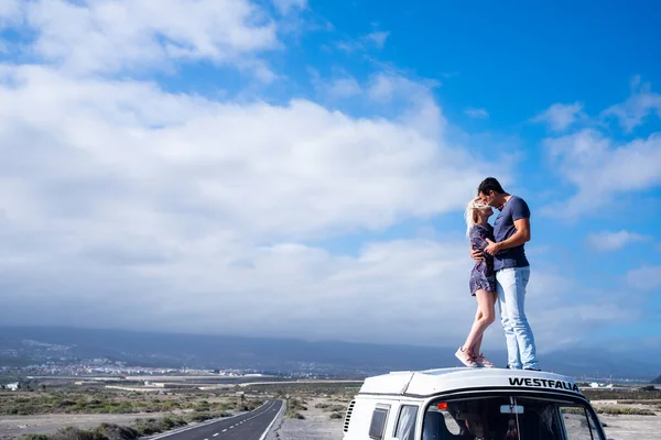 Love and travel people lifestyle, standing couple on the roof vehicle van together kissing and hugging with long road and blue sky in background