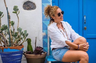 Güzel bir kadın evinin önünde oturur ve rahatlar. Rahatlama içinde, rahat kadın aktivitesi arka planda mavi kapı ve ev ile dışarıda.