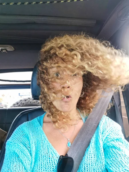 Surprised face expression adult beautiful woman inside the car with safety belt and wind moving the curly hair