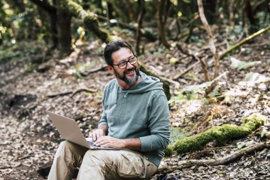 Mutlu yetişkin adam yere otur ve dizüstü bilgisayarla internet bağlantısıyla çalış. Kablosuz kablosuz kablosuz internetin arka planında orman odunu var.