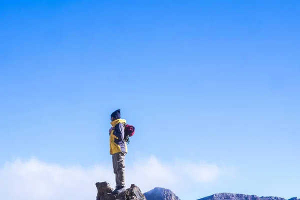 冒险家 穿着暖和的衣服 头戴针织的帽子 背着背包 站在多石的山顶上 蓝云的天空映衬着他 — 图库照片
