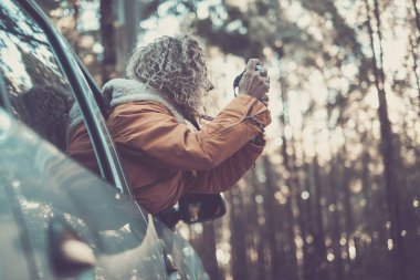 Sıcak ceketli bir kadın arabanın camından sarkıyor. Orman boyunca kamera kullanarak fotoğraf çekiyor.