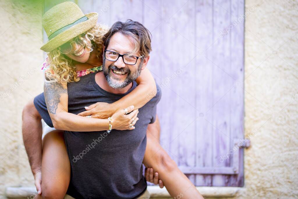 Carefree couple spending leisure time together outdoors, man giving piggyback ride to cheerful tattooed woman in sunglasses and straw hat. 