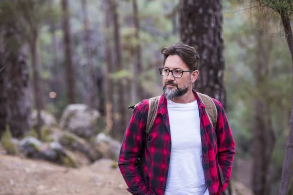 Thoughtful handsome man in eyeglasses and checked shirt looking away while admiring something interesting on a hiking tour in forest.