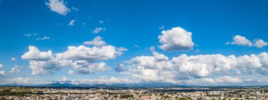 Image of a day with the blue sky and some white clouds. At the base of the image is a city and mountains in the background. 
