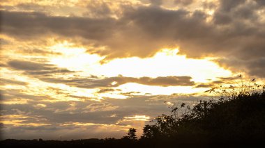 Gün batımı turuncu renkle vurgulanmış, dramatik bulutlar, ufkun altında ve sağ köşede bir ağacın silueti