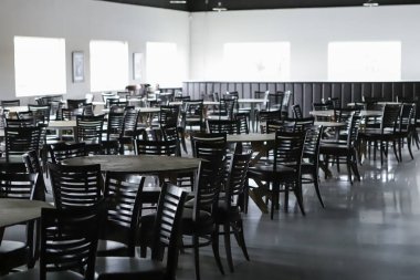 Boş balo salonu, birkaç yuvarlak masa, dekorasyon yok, ve koyu kahverengi sandalyeler. Arka planda ışık neredeyse patlamak üzere olan pencereler..