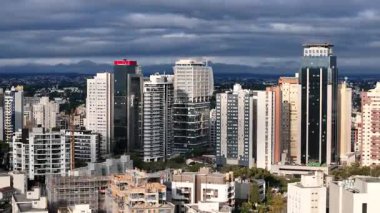 Curitiba şehrinin, kısmen bulutlu bir gökyüzünün altında, çok sayıda modern bina ve gökdelenle dolu yoğun bir kentsel alandaki hava görüntüsü. Arka planda, daha fazla bitki örtüsü ve tepeleri olan bir manzara.
