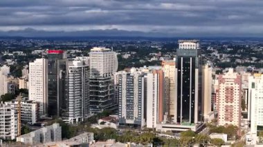 Curitiba şehrinin, kısmen bulutlu bir gökyüzünün altında, çok sayıda modern bina ve gökdelenle dolu yoğun bir kentsel alandaki hava görüntüsü. Arka planda, daha fazla bitki örtüsü ve tepeleri olan bir manzara.