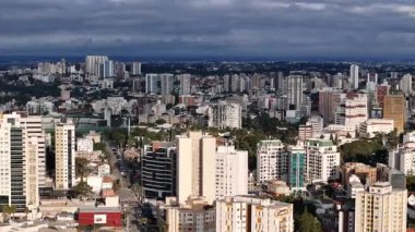Curitiba şehrinin, kısmen bulutlu bir gökyüzünün altında, çok sayıda modern bina ve gökdelenle dolu yoğun bir kentsel alandaki hava görüntüsü. Arka planda, daha fazla bitki örtüsü ve tepeleri olan bir manzara.