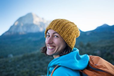 Sırt çantasıyla dağların arkasında gülümseyen bir kızın portresi. Türkiye 'de seyahat. Örgü şapkalı bir kadın bir dağın tepesinde duruyor..