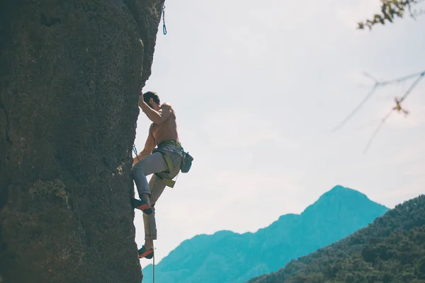 A rock climber climbs a rock against the background of mountains and sky, a man trains strength and endurance, overcoming the fear of heights, training in nature.