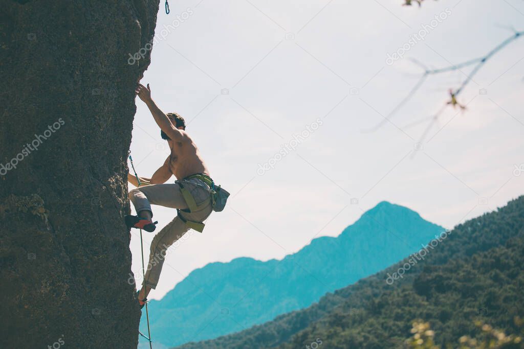 A rock climber climbs a rock against the background of mountains and sky, a man trains strength and endurance, overcoming the fear of heights, training in nature.