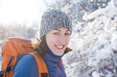 Örgü şapkalı bir kızın portresi. Karlı bir ormanın arka planında sırt çantalı gülümseyen bir kadın. Kış yürüyüşüne çıkmış bir kadın. Kış yükseliyor dağlara.