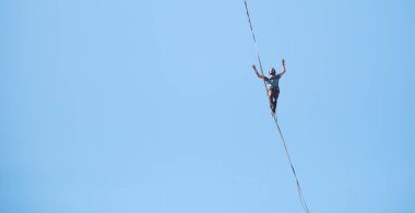 Bir adam mavi gökyüzüne karşı Highline boyunca yürüyor. Highliner askı takıyor. İp cambazı. Bir adam dengeyi yakalar..