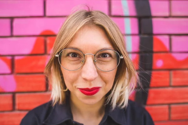 Portrait of a blonde in glasses on a brick wall background. A woman walks around the city. The smiling girl. Lips with red lipstick.