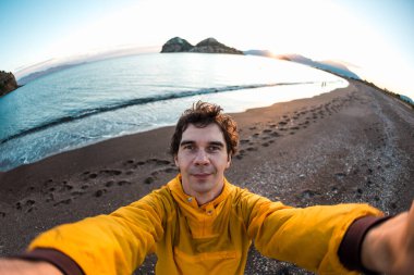 Sarı ceketli genç adam gün batımında deniz kıyısında selfie çekiyor. Türkiye 'ye gitmek. Sadece rahatlama ve macera..