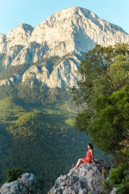 Kırmızı elbiseli bir kız dağın tepesindeki büyük bir kayanın tepesinde bir dağ vadisinin arka planında oturuyor, Türkiye 'nin pitoresk yerlerine bir gezi..