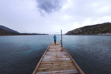 Bir kız rıhtımda duruyor ve denize bakıyor, bir kadın rıhtımın ortasında duruyor, pitoresk yerlere bir gezi, gölün arka planında bir kız, dağlar ve bulutlu bir gökyüzü, soğuk mevsim