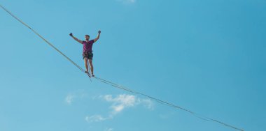 Mavi gökyüzünün arka planındaki Highliner bir hamle yapıyor. Bir adam askı manzarası boyunca aşağıdan yürüyor. Performans ip cambazı. İnsanoğlu uçurumun üzerinde denge kurar. İleriye doğru kararlı bir adım.