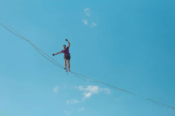 在蓝天的背景下 高耸的轮廓使一个动作 一个男人从下面沿着一条伸展的吊带的景色走着 表演走钢丝 人在深渊中保持平衡 向前迈出决定性的一步 — 图库照片