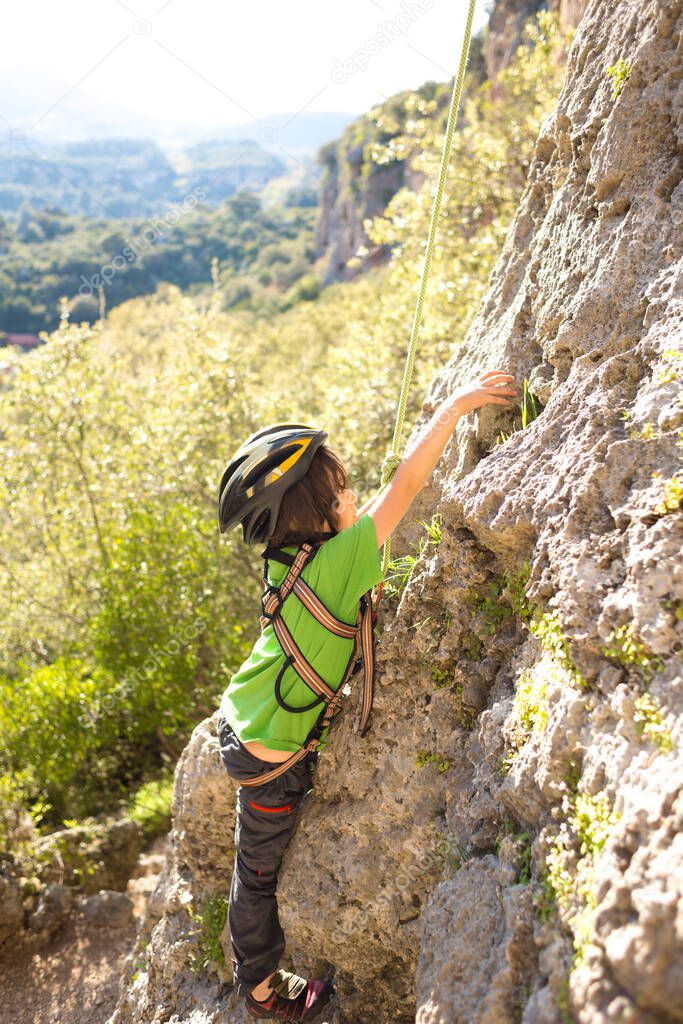 The child is climbing on a natural terrain. A boy in helmet climbs a rock on a background of mountains. Extreme hobby. Athletic kid trains to be strong. Rock climbing safety.
