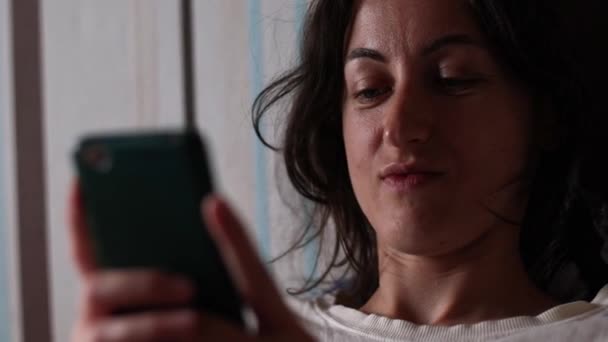 微笑的女孩躺在沙发上 在智能手机上留言 女人在休息 黑发女人读书微笑 — 图库视频影像