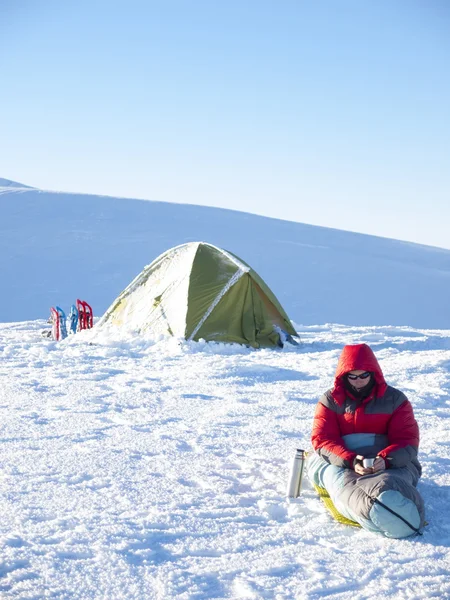 Ein Mann sitzt in einem Schlafsack neben dem Zelt und Schneeschuhen. — Stockfoto