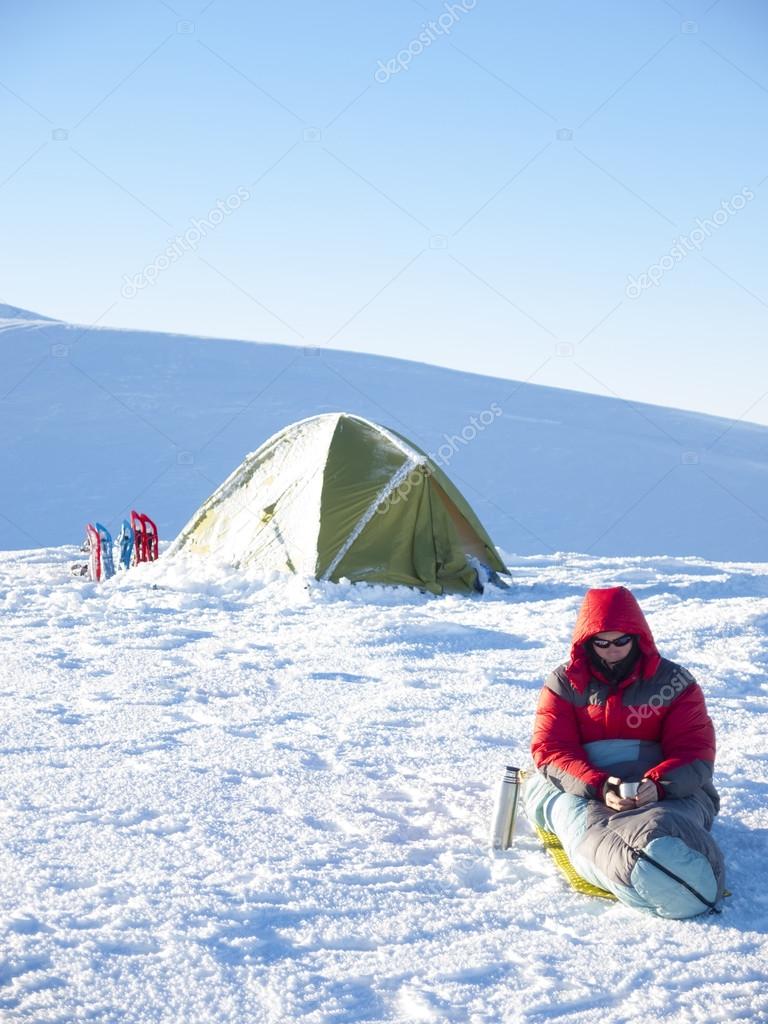 A man sits in a sleeping bag near the tent and snowshoes.