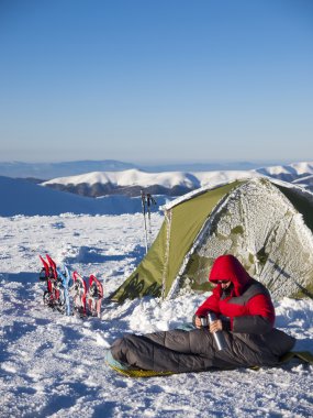 Çadır ve kar ayakkabıları yakınındaki uyku tulumu içinde bir adam oturuyor.