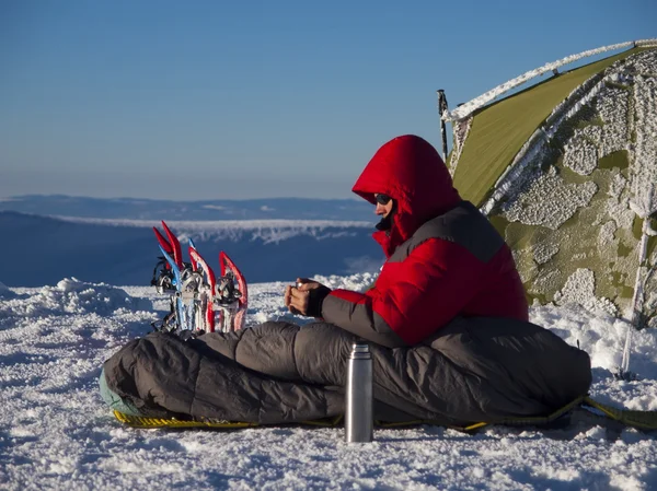 En man sitter i en sovsäck nära tält och snöskor. — Stockfoto