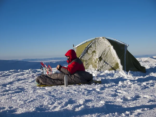 Człowiek siedzi w śpiwór w pobliżu namiotu i rakiety śnieżne. — Zdjęcie stockowe