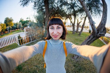 Sırt çantalı, selfie çeken ve yaz parkında yürürken kameraya bakan pozitif bir kız resmi. Kız seyahat eder.